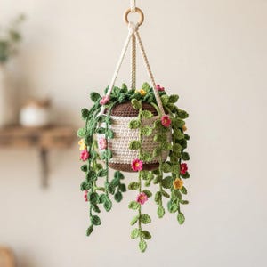 May include: A crocheted hanging plant in a two-tone brown and cream pot. The plant features long, trailing green vines with small, detailed leaves and tiny pink and yellow flowers. The entire piece is suspended by a cream-colored cord and a wooden ring.