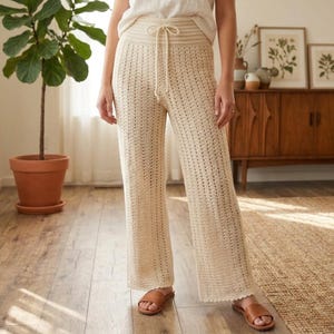 May include: Cream-colored crochet pants featuring a high-waisted design with a drawstring closure. The trousers are crafted with a vertical open-knit pattern, a wide-leg silhouette, and a scalloped hemline, providing a textured look.