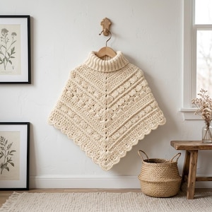 May include: A cream-colored crochet poncho featuring a ribbed turtleneck collar and a textured pattern of raised shell stitches arranged in a V-shape. The garment has a scalloped hemline and is displayed hanging on a wooden hanger against a wall.