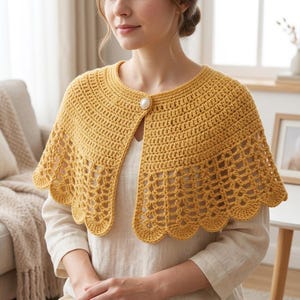 Puede incluir: Una esclavina de ganchillo color amarillo mostaza con un patrón de puntos densos y sólidos en el cuello y un borde festoneado calado decorativo a lo largo del dobladillo. La prenda se sujeta en el centro delantero con un botón redondo nacarado.