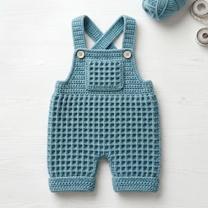 Puede incluir: Un peto de ganchillo azul con un patrón de punto de gofre en todo el cuerpo y un bolsillo cuadrado en el pecho. La prenda incluye dos tirantes que se cruzan en la espalda y se aseguran con dos botones de madera clara en la parte delantera.