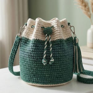 May include: A crochet bucket bag featuring a dark green base and a cream-coloured top section. The bag includes a matching dark green crochet shoulder strap, a drawstring closure with twisted cream and green cords, and a decorative tassel.