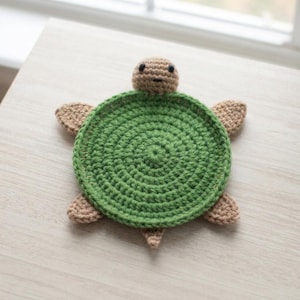 May include: A crochet turtle coaster featuring a circular green shell with a textured stitch pattern. The turtle has a light brown head with two small black bead eyes, four matching light brown flippers, and a small tail extending from the shell.