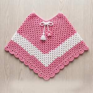 May include: A pink and white crochet poncho featuring a V-shaped pattern with a scalloped edge. The garment includes a white drawstring tie at the neckline, finished with a small white tassel and a matching pink crocheted flower embellishment.