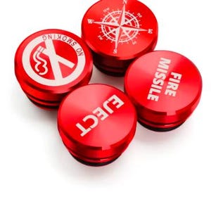 Op de afbeelding: Vier ronde vervangende sigarettenaanstekerpluggen van rood metaal. Elk exemplaar heeft een unieke witte afbeelding of tekst: een kompasroos, een rookverbodsymbool, het woord EJECT en de tekst FIRE MISSILE. De knoppen hebben een gladde afwerking.