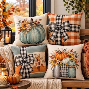 May include: Four square throw pillows featuring autumn themes. Designs include a teal pumpkin with sunflowers, an orange buffalo check pattern with a black and white bow, a teal and orange striped pillow with a bow, and pumpkins in jars.
