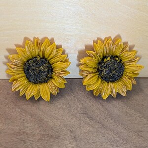 May include: Two sunflower-shaped objects featuring bright yellow petals radiating from a dark, textured brown center. The petals exhibit detailed ridges and depth, creating a realistic floral appearance with a glossy finish on the surface.