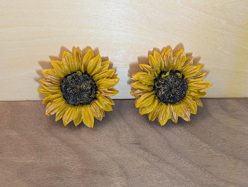May include: Two sunflower-shaped objects featuring bright yellow petals radiating from a dark, textured brown center. The petals exhibit detailed ridges and depth, creating a realistic floral appearance with a glossy finish on the surface.