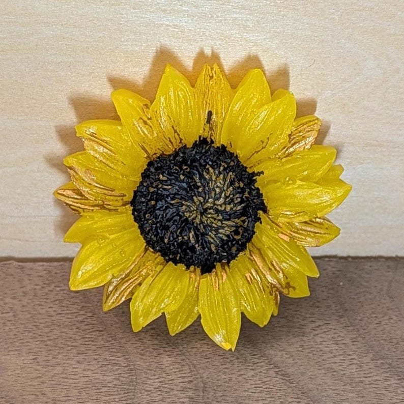May include: A sunflower-shaped decorative object featuring bright yellow petals with subtle gold-toned highlights and a textured, dark brown center. The piece displays a realistic floral design with layered, pointed petals radiating from the core.