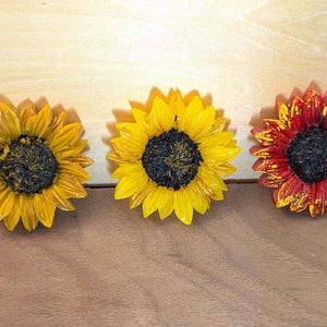 May include: Three decorative sunflower ornaments arranged in a row. The left flower is golden yellow, the center flower is bright yellow, and the right flower features a mix of deep red and yellow petals. Each has a dark, textured center.