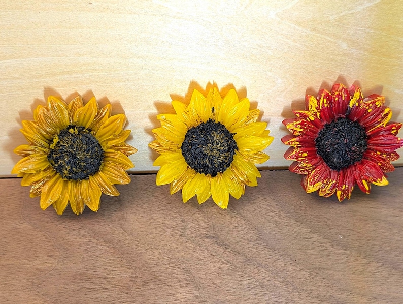 May include: Three decorative sunflower ornaments arranged in a row. The left flower is golden yellow, the center flower is bright yellow, and the right flower features a mix of deep red and yellow petals. Each has a dark, textured center.