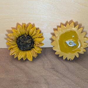 May include: Two sunflower-shaped objects, one showing the front with yellow petals and a dark brown center, and the other showing the back with a smooth yellow surface and a small silver-colored metal attachment point in the center.