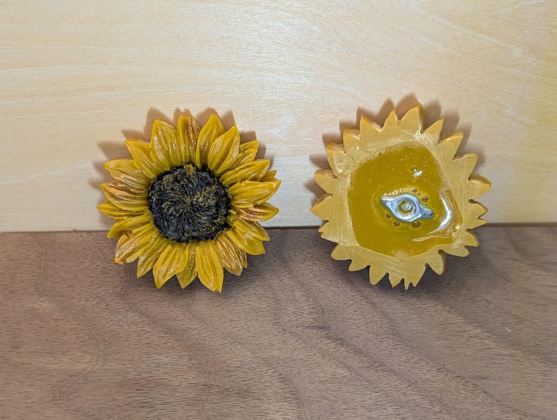 May include: Two sunflower-shaped objects, one showing the front with yellow petals and a dark brown center, and the other showing the back with a smooth yellow surface and a small silver-colored metal attachment point in the center.
