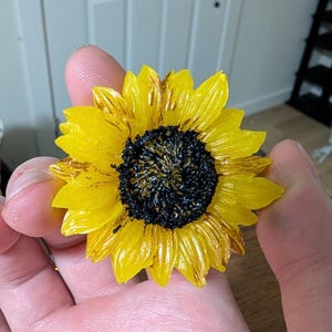 May include: A sunflower-shaped object with bright yellow petals featuring subtle brown streaks and a textured, dark black center. The flower has a layered, realistic appearance with defined petal shapes radiating from the central disc.