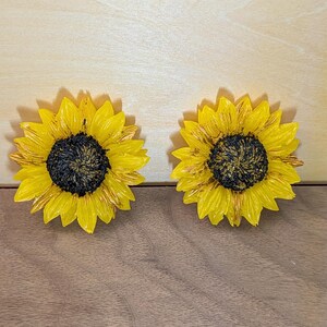 May include: Two sunflower-shaped decorative items featuring bright yellow petals with subtle brown highlights and a textured, dark brown center. The sunflowers are round with layered, pointed petals radiating from the center, creating a realistic floral appearance.