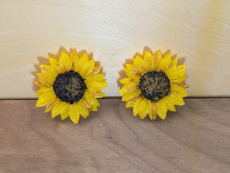 May include: Two sunflower-shaped decorative items featuring bright yellow petals with subtle brown highlights and a textured, dark brown center. The sunflowers are round with layered, pointed petals radiating from the center, creating a realistic floral appearance.