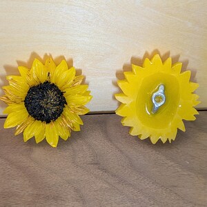 May include: Two sunflower-shaped decorative items. The left item shows the front with bright yellow petals and a dark brown textured center. The right item shows the back, which is yellow and features a silver-colored metal attachment hardware.