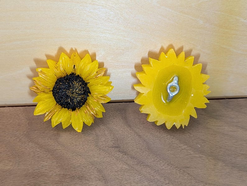 May include: Two sunflower-shaped decorative items. The left item shows the front with bright yellow petals and a dark brown textured center. The right item shows the back, which is yellow and features a silver-colored metal attachment hardware.