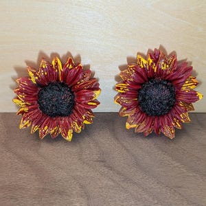 May include: Two sunflower-shaped decorative items featuring deep burgundy red petals with bright yellow tips and a textured, dark brown center. The petals exhibit a layered, realistic appearance with a glossy finish, showcasing a distinct contrast between the dark red and yellow colors on each flower.