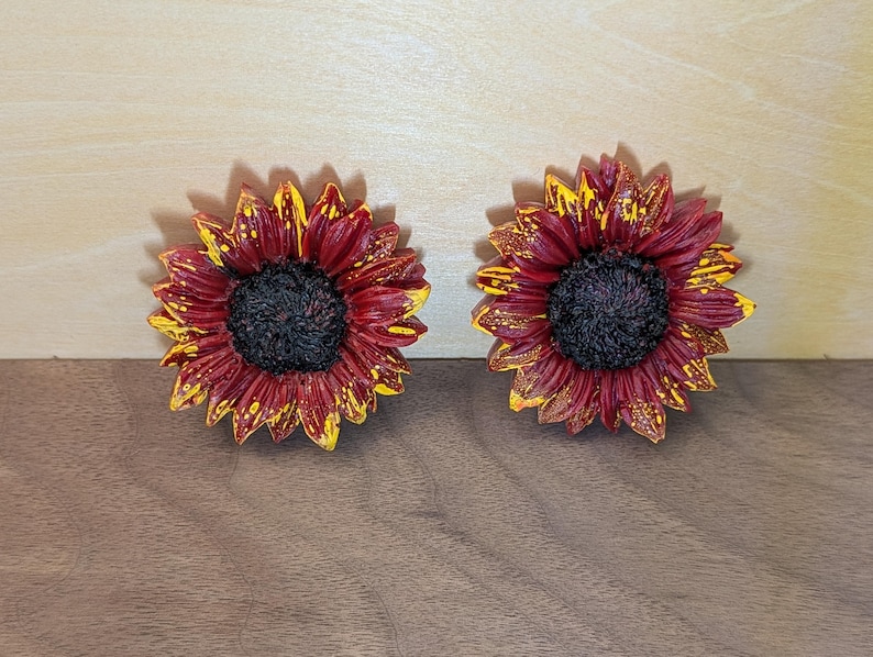 May include: Two sunflower-shaped decorative items featuring deep burgundy red petals with bright yellow tips and a textured, dark brown center. The petals exhibit a layered, realistic appearance with a glossy finish, showcasing a distinct contrast between the dark red and yellow colors on each flower.