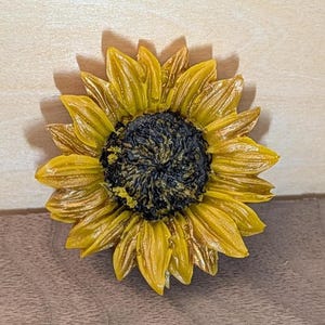 May include: A sunflower-shaped object featuring a dark, textured center surrounded by multiple layers of pointed, golden-yellow petals. The petals exhibit a glossy finish with subtle variations in tone, radiating outward from the central disc.