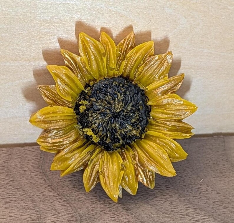 May include: A sunflower-shaped object featuring a dark, textured center surrounded by multiple layers of pointed, golden-yellow petals. The petals exhibit a glossy finish with subtle variations in tone, radiating outward from the central disc.