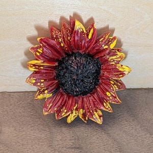 May include: A decorative sunflower ornament featuring deep red petals with bright yellow tips and speckles. The center of the flower is textured and dark black, creating a high-contrast appearance. The petals are layered and radiate outward from the center.
