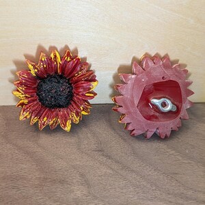 May include: Two sunflower-shaped decorative pieces. One shows the front with deep red petals tipped in yellow and a dark textured center. The other shows the back, which is solid red with a central metal attachment point featuring a small screw hole.