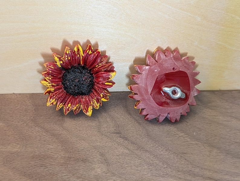 May include: Two sunflower-shaped decorative pieces. One shows the front with deep red petals tipped in yellow and a dark textured center. The other shows the back, which is solid red with a central metal attachment point featuring a small screw hole.