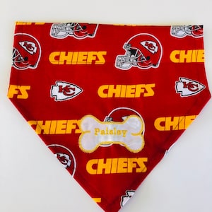 May include: Red and yellow dog bandana with a white bone-shaped tag that says "Paisley" and a pattern of football helmets and the word "Chiefs".