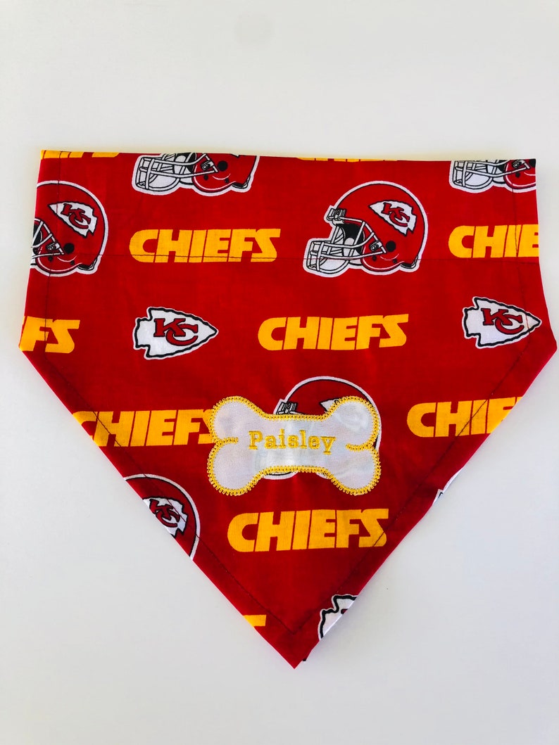 May include: Red and yellow dog bandana with a white bone-shaped tag that says "Paisley" and a pattern of football helmets and the word "Chiefs".