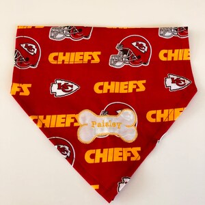 May include: Red and gold dog bandana with a white bone-shaped tag that says "Paisley" and a pattern of Kansas City Chiefs logos.