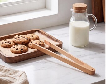 Op de afbeelding: Een lichtgekleurde houten keukentang met een geribbeld uiteinde, geplaatst naast een houten dienblad met vier ronde koekjes met chocoladestukjes en een glazen karaf gevuld met melk, afgesloten met een ronde kurken stop.