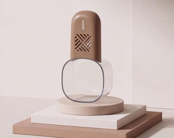 Op de afbeelding: Een bruin kunststof keukengereedschap met een afgeronde bovenkant, een ventilatieopening met een rasterpatroon en het woord "spoon" verticaal gedrukt. Het bovenste gedeelte is bevestigd aan een doorzichtige, vierkante houder.
