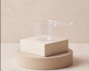 Op de afbeelding: Een doorzichtige plastic maatschep met een cilindervormig lichaam en een plat handvat aan de zijkant. De buitenkant van de maatschep is voorzien van horizontale lijnen om volumeniveaus aan te geven voor het afmeten van diverse ingrediënten.