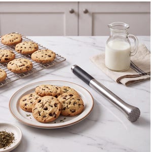 Puede incluir: Un mortero de acero inoxidable con mango negro colocado junto a un plato blanco que contiene cuatro galletas con chispas de chocolate, una rejilla de enfriamiento con más galletas y una pequeña jarra de vidrio con leche.