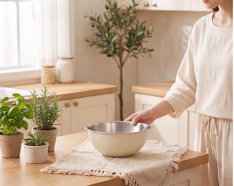 Op de afbeelding: Een ronde mengkom met een crèmekleurige buitenkant en een gepolijste roestvrijstalen binnenkant. De kom staat op een vierkant, beige stoffen servet met franjes. De rand van de kom is glad en metaalachtig, wat contrasteert met de matte afwerking.