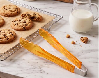 Op de afbeelding: Een keukentang met doorschijnende oranje kunststof handgrepen met geschulpte randen en een roestvrijstalen basis. De tang ligt naast een afkoelrek met chocoladekoekjes en een glazen kan met melk.