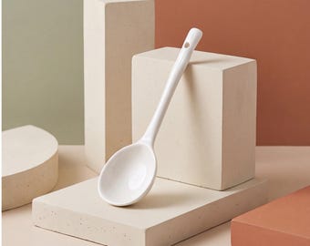 Op de afbeelding: Een glanzende witte keramische lepel met een gladde, ronde kom en een lange, slanke steel. De steel is voorzien van een klein, rond gat aan het uiteinde, bedoeld om de lepel op te hangen. De lepel heeft een egale, volledig witte afwerking.