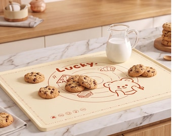 Op de afbeelding: Een rechthoekige, crèmekleurige siliconen bakmat met bruine lijntekeningen van een beer, het woord Lucky en diverse voedselpictogrammen. De mat bevat cirkelvormige meetgidsen en een rand met liniaalmarkeringen, met daarop chocoladekoekjes.