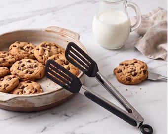 Op de afbeelding: Een keukentang met zwarte siliconen uiteinden, roestvrijstalen handgrepen en een ronde ophanglus, geplaatst naast een keramische schaal gevuld met chocoladekoekjes en een glazen kan met witte melk.