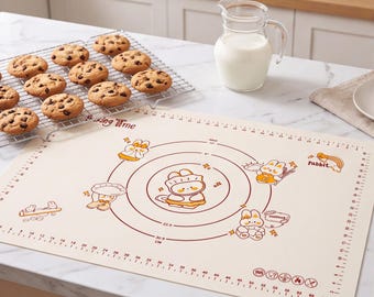 Op de afbeelding: Een crèmekleurige siliconen bakmat met een konijnenthema, concentrische cirkels, maataanduidingen in centimeters en de tekst "Baking Time" en "rabbit". Een taartrooster met chocoladekoekjes staat ernaast.