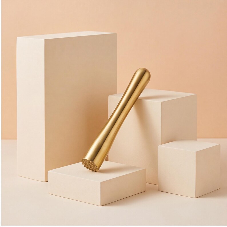Puede incluir: Un mortero para c&oacute;cteles de metal dorado con un mango cil&iacute;ndrico liso y una base texturizada y dentada. La herramienta est&aacute; colocada en &aacute;ngulo, apoyada contra una serie de bloques geom&eacute;tricos de color beige claro de diversas alturas.