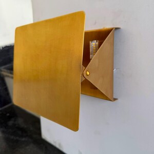 May include: A wall-mounted light fixture featuring a rectangular brass-toned metal plate attached to a triangular bracket. The fixture includes an exposed LED component visible within the side opening of the metal structure.