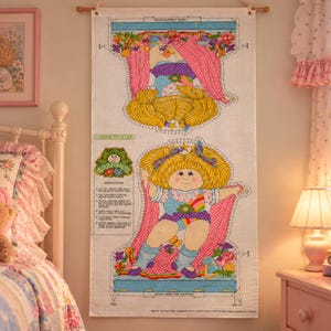 Op de afbeelding: Een Cabbage Patch Kids stoffen paneel met een bedrukt poppenontwerp met geel haar, een blauw-wit gestippelde top en een regenboogafbeelding. Het paneel bevat naai-instructies en kniplijnen voor het maken van een stoffen pop en kleding.