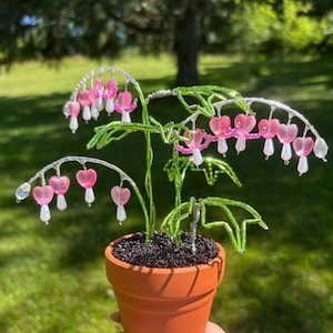 Op de afbeelding: Een decoratieve plantsculptuur gemaakt van draad en kralen, met roze hartvormige bloesems en groene bladvormige kralen. De stengels zijn gemaakt van gedraaid zilverdraad en de plant staat in een kleine terracotta-kleurige aarden pot.