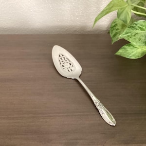 Puede incluir: Un servidor de pasteles de metal plateado con una hoja triangular puntiaguda que presenta un intrincado diseño calado en el centro y un mango delgado decorado con patrones florales en relieve en los bordes.