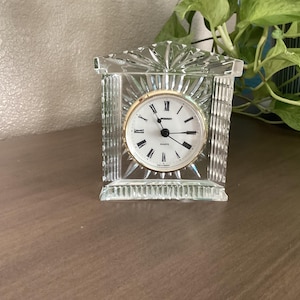 May include: A clear glass mantel clock with a rectangular, faceted frame and a triangular top. The clock face is white with black Roman numerals, black hands, and a gold-tone bezel. The text on the face reads Quartz and Made in Germany.