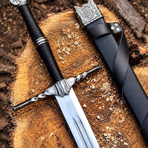 Op de afbeelding: Een stalen zwaard met een zwart omwikkeld handvat en een zilveren pommel met een wolfskopembleem. De pareerstang heeft een gedraaid metalen ontwerp. Een bijpassende zwarte lederen schede met een versierd zilveren mondstuk ligt ernaast.