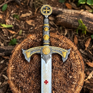 Puede incluir: Una espada decorativa con empuñadura plateada y dorada con grabados intrincados, un pomo circular con una cruz y una guarda con detalles dorados. La hoja muestra adornos ornamentales y un símbolo de cruz roja cerca de la empuñadura.